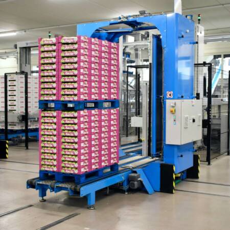„Automatisierte Verpackungsanlage mit einer großen blauen Maschine, die palettierte Waren in einer modernen Produktionshalle verarbeitet.“