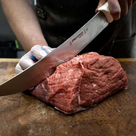 „Metzger schneidet ein großes Stück rohes Rindfleisch mit einem scharfen Messer auf einem Holzbrett, trägt dabei Handschuhe für Hygiene.“